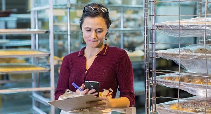 Woman in bakery looking at phone and clipboard